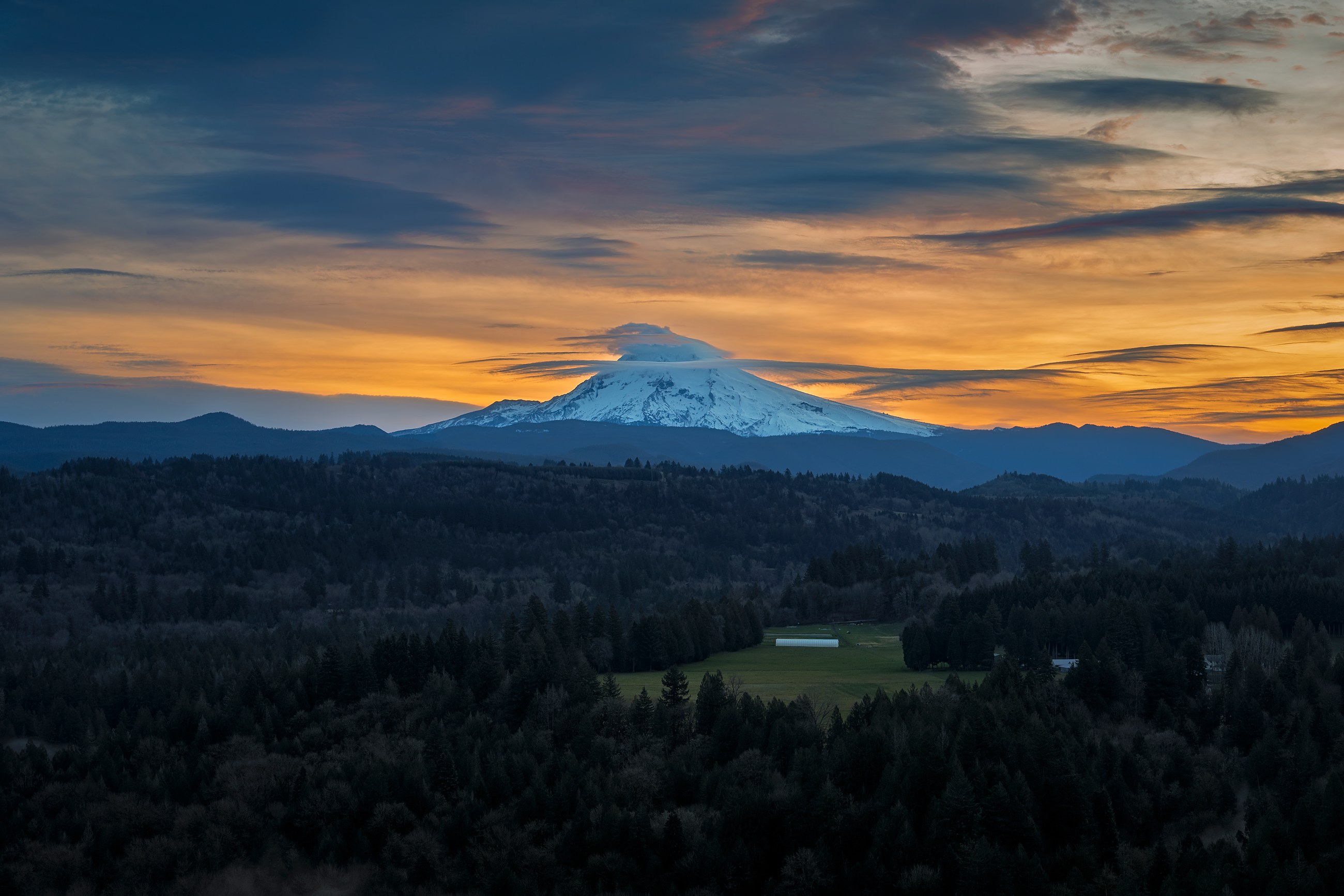 Mt Hood with a cloud cap at sunset, orange sky over the forested valley below, Oregon Cascades